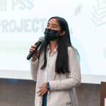 Young adult standing in front of a screen speaks into a microphone